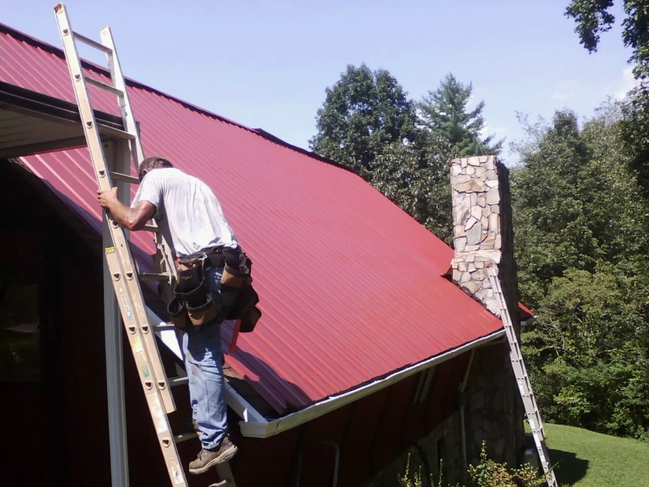 Roofer on a ladder beside a red metal roof and stone chimney — Soffit & Fascia Repair by Roofing Brothers in Whitwell